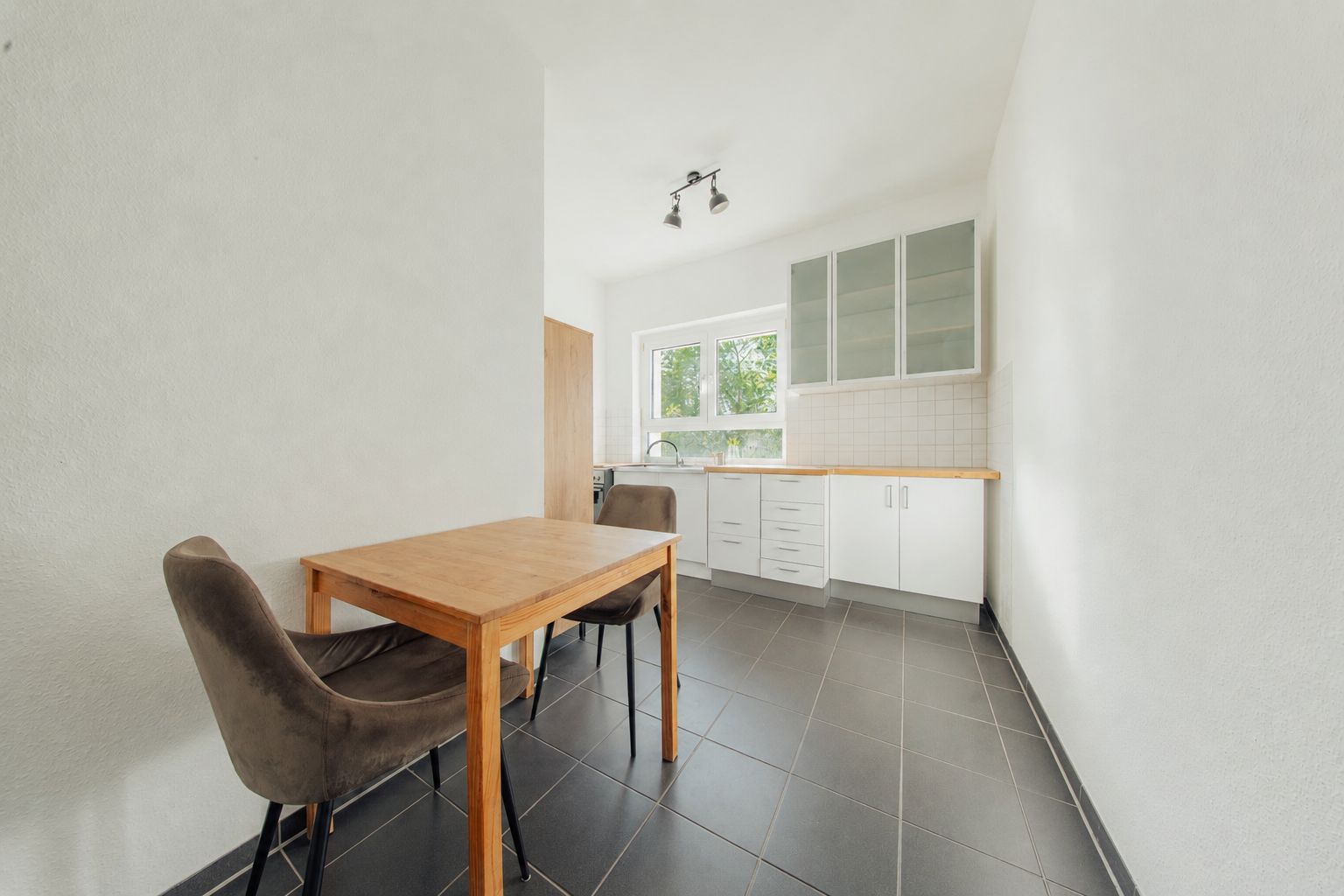 Helle Küche mit Esstisch, zwei Stühlen, weißen Schränken und großem Fenster.