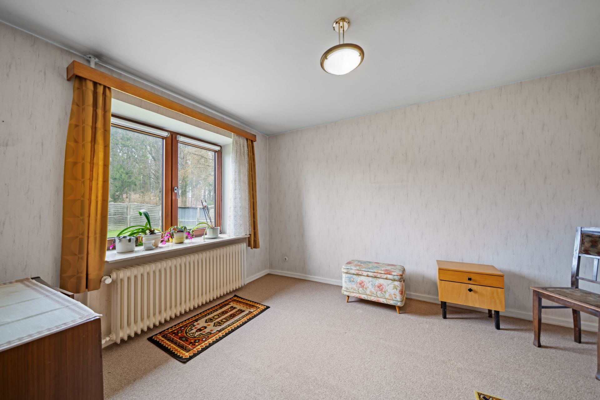 Helles Zimmer mit großem Fenster, gelben Vorhängen, Teppich, Kommode und Hocker.