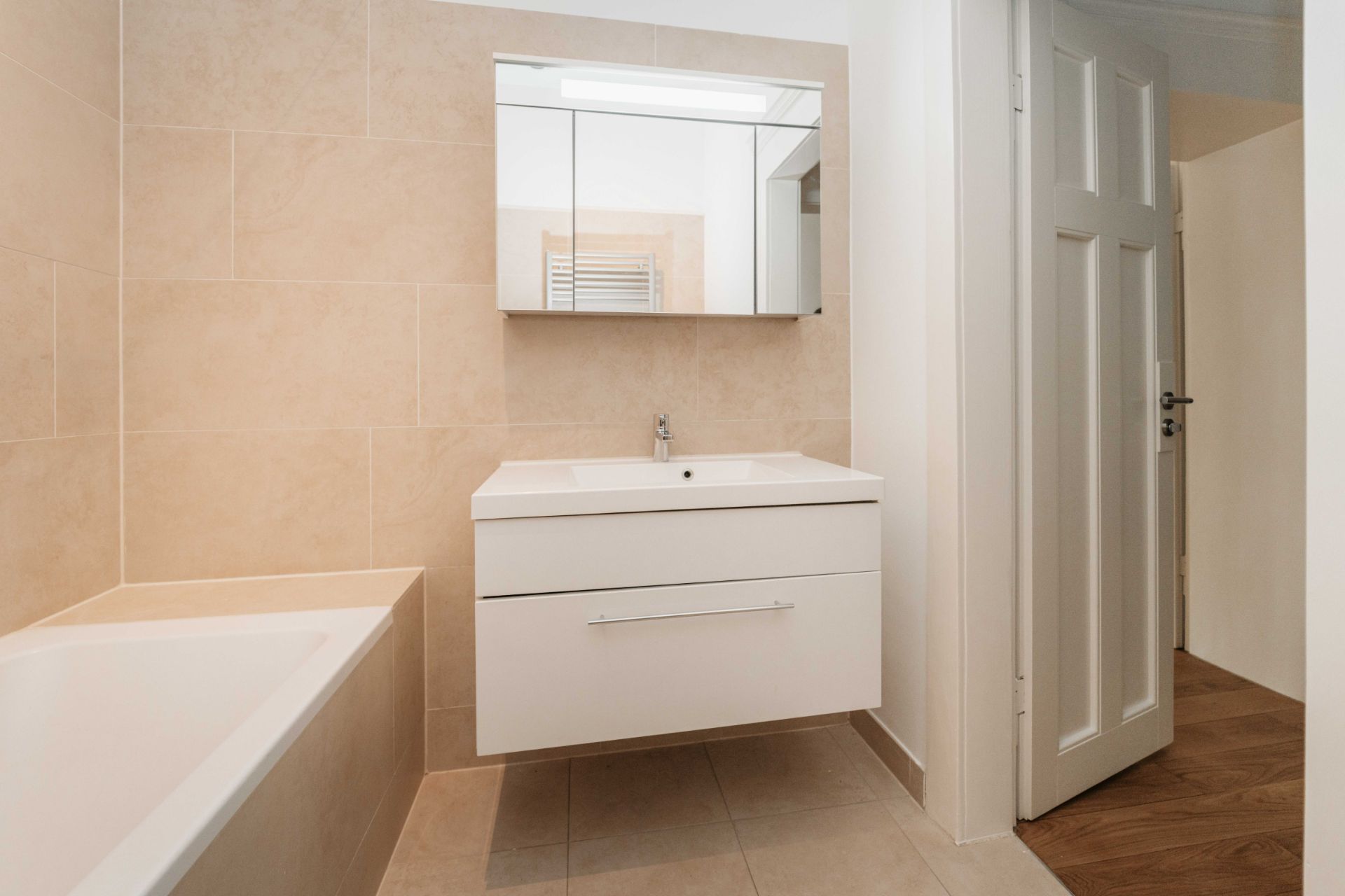 Modernes Bad mit beiger Fliesen, Badewanne, weißem Waschbecken und Spiegelschrank.
