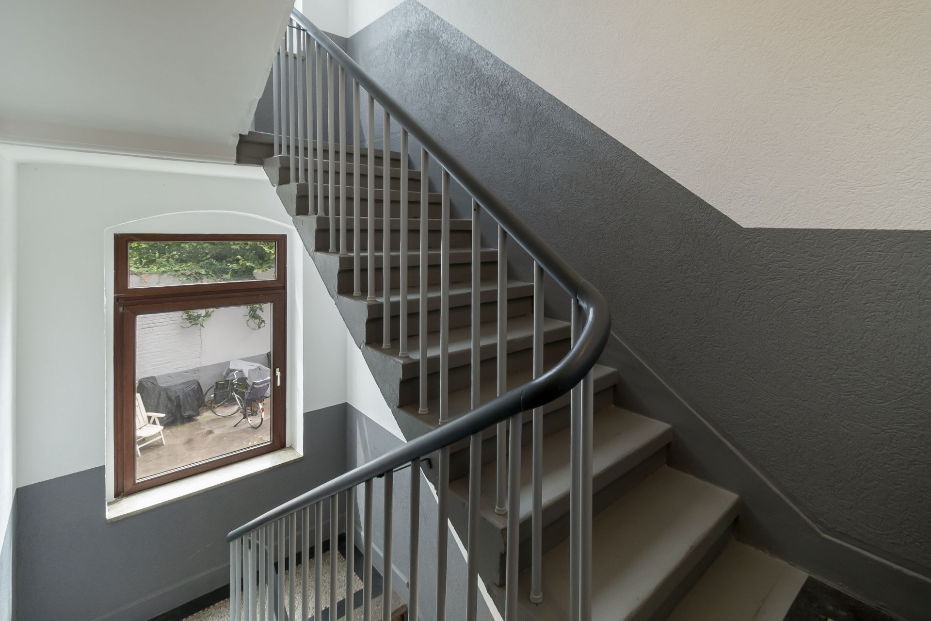 Treppe mit grauem Geländer und Fenster, das auf einen Innenhof mit Fahrrad blickt.