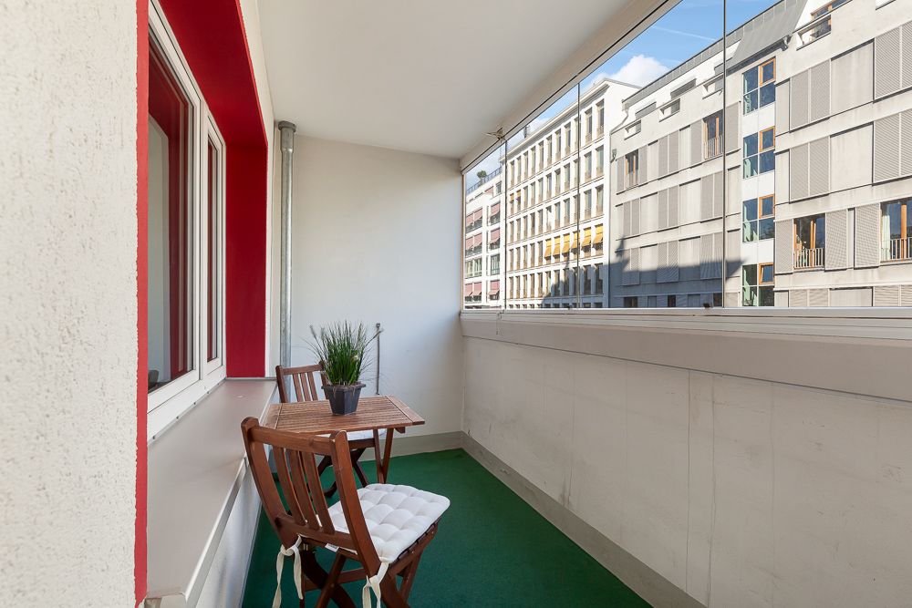 Überdachter Balkon mit zwei Holzstühlen, kleinem Tisch und Pflanze, Blick auf Nachbargebäude.