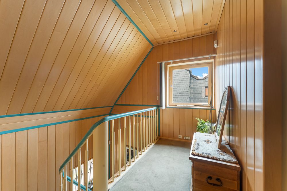 Heller Flur mit Holzvertäfelung, Fenster, Holzgeländer und Kommode unter schräger Decke.