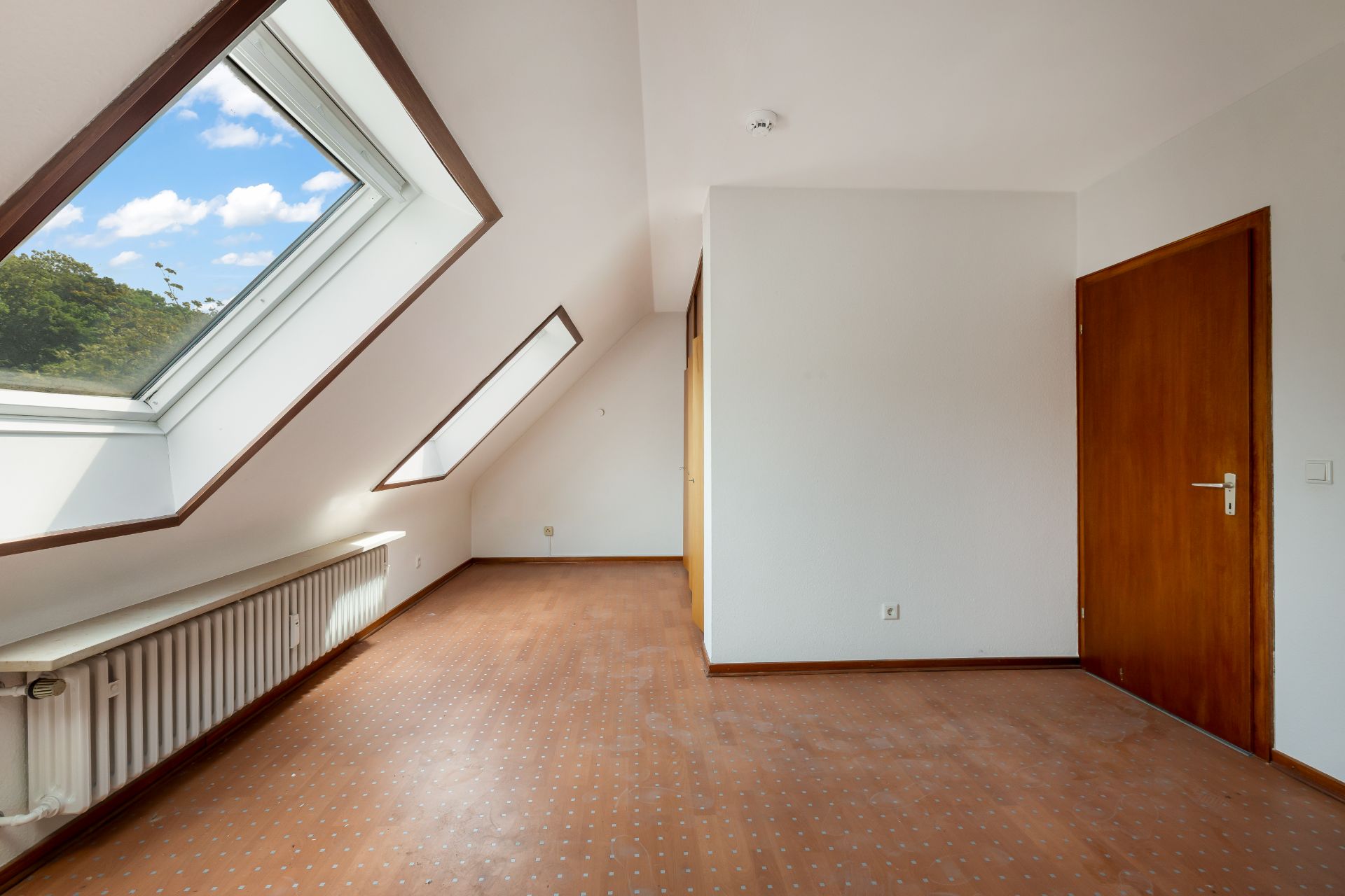 Heller Dachzimmer mit schräger Decke, zwei großen Dachfenstern und Holztür.