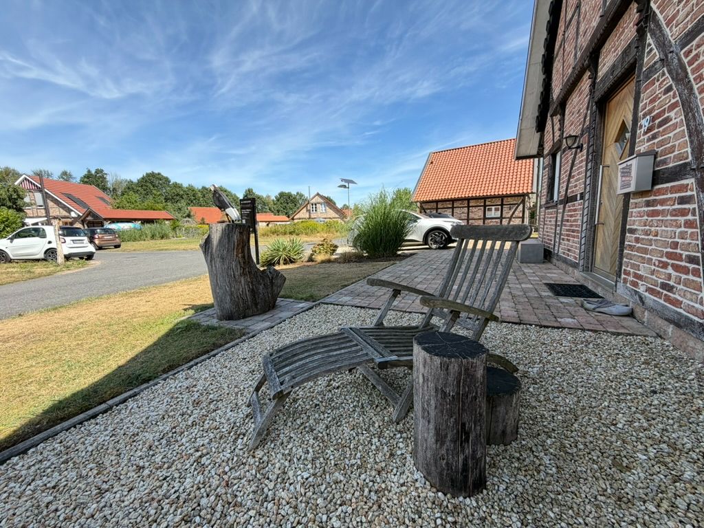 Holzstuhl und Tisch auf Kiesfläche vor einem Fachwerkhaus mit Garten und parkenden Autos.