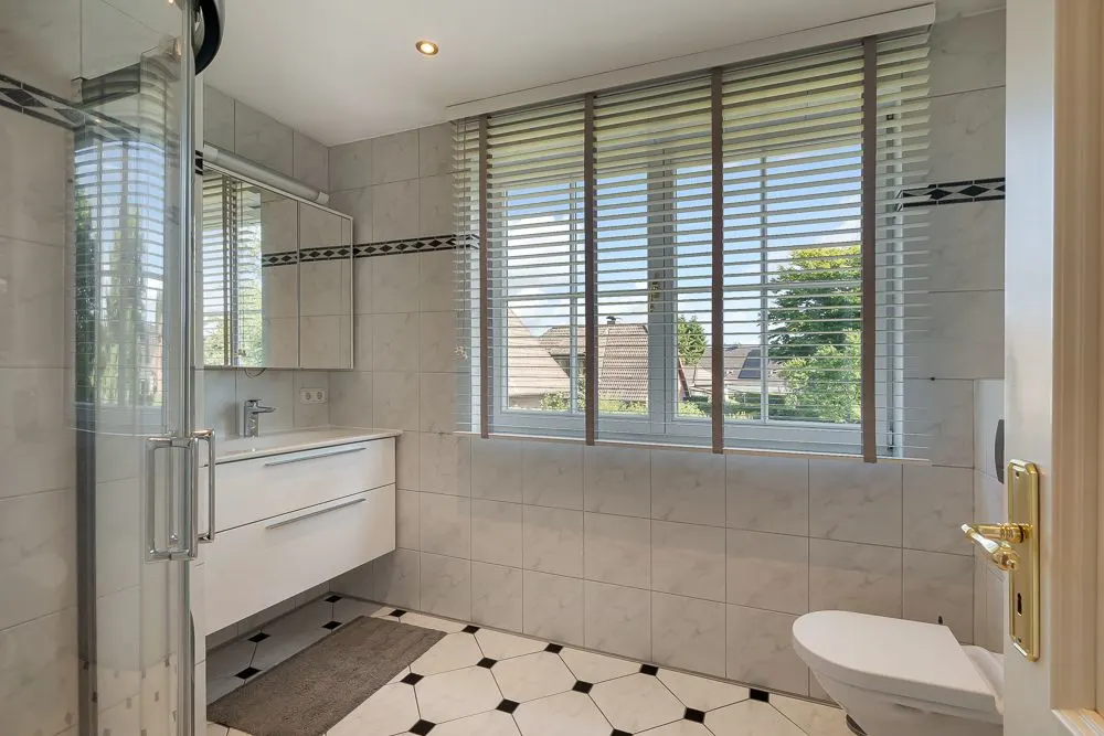Helles Badezimmer mit großer Fensterfront, Dusche, Waschbecken und WC.