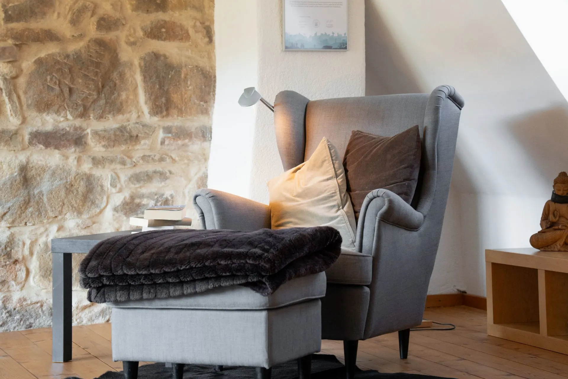 Grauer Sessel mit Hocker, Kissen und Decke neben einem Beistelltisch vor einer Steinwand.