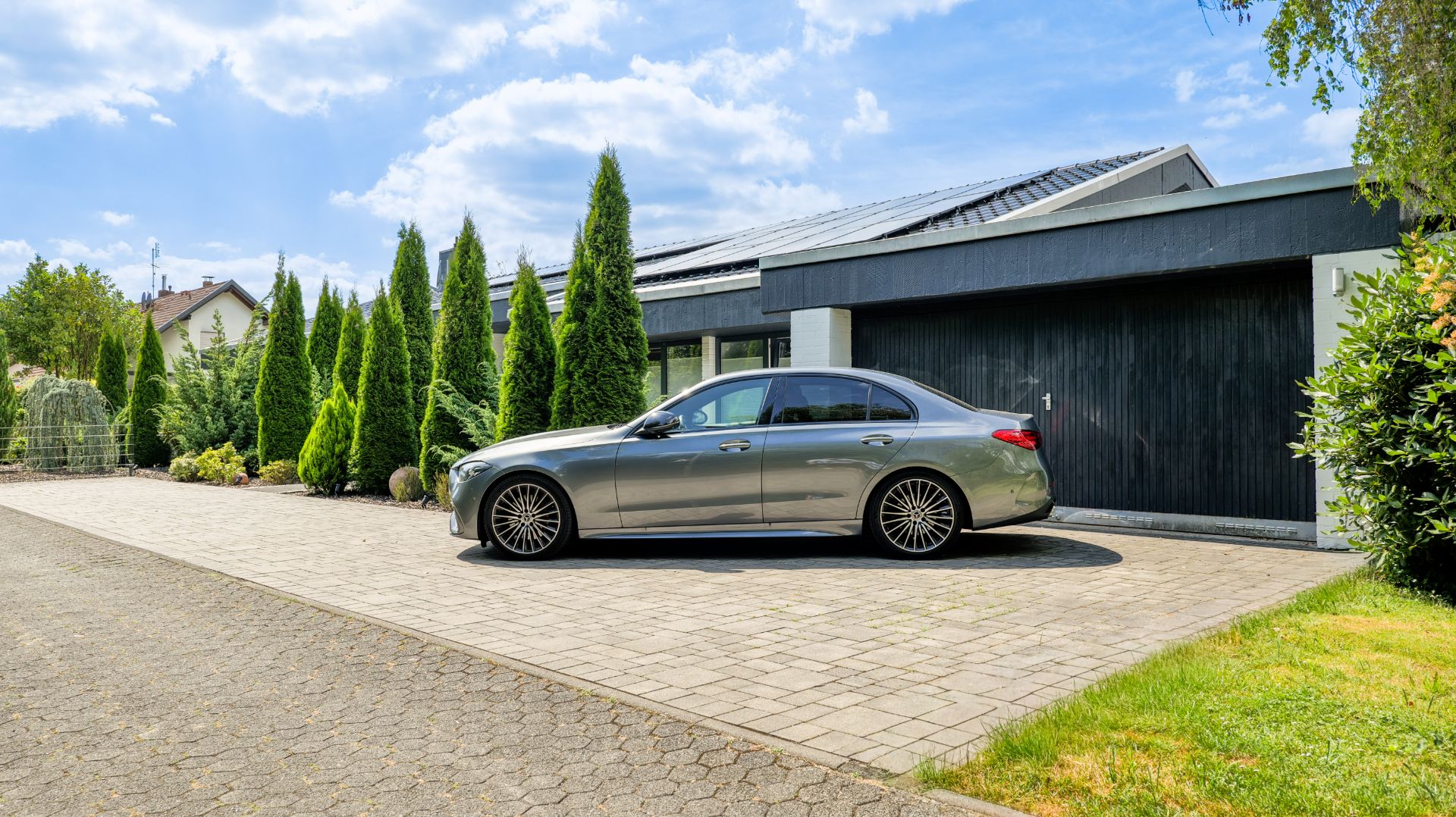 Silbernes Auto parkt vor einer modernen Garage mit hohen grünen Bäumen daneben.