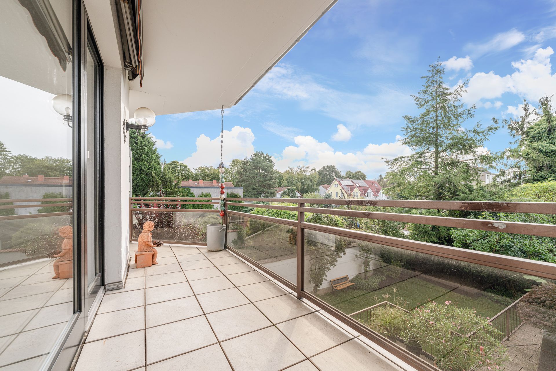 Großer Balkon mit Glasgeländer, Blick auf Garten und Bäume, Statue und Blumentopf auf den Fliesen.