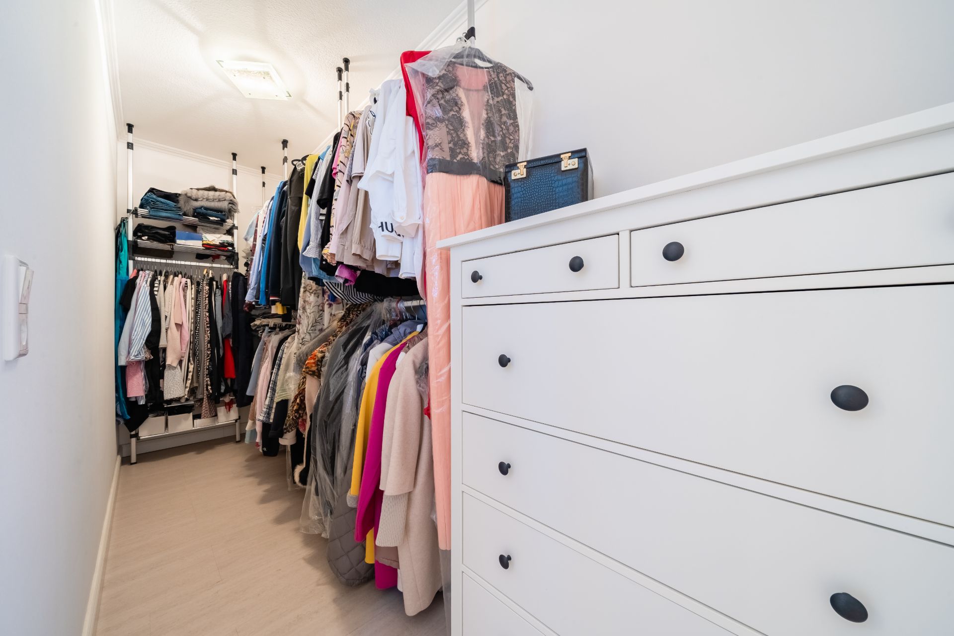 Begehbarer Kleiderschrank mit weißer Kommode und vielen Kleidungsstücken auf Kleiderstangen.