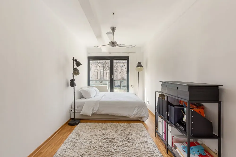 Schlafzimmer mit Doppelbett, Steh- und Leselampe, Teppich und Regal, großes Fenster im Hintergrund.
