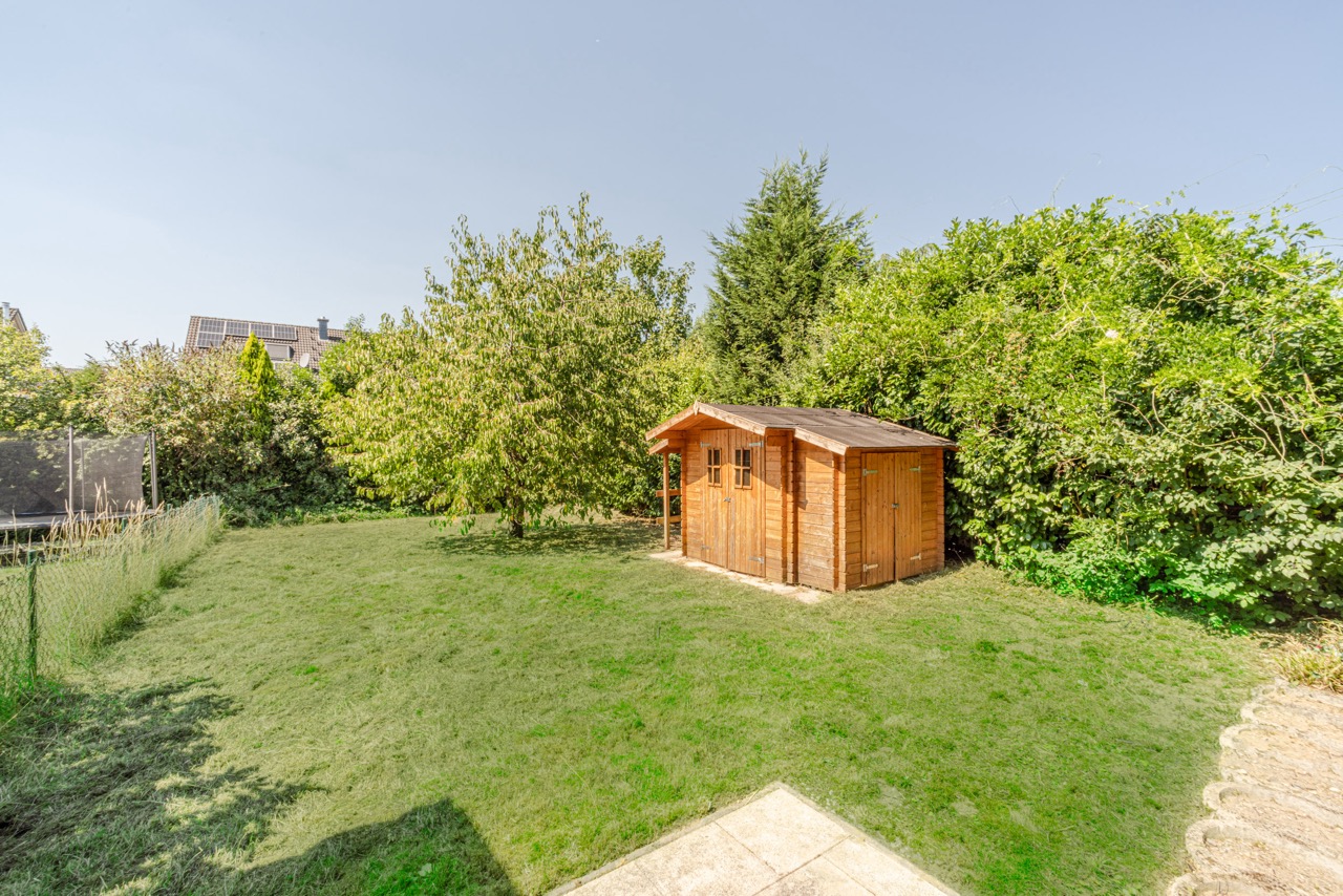 Garten mit Rasenfläche, Holzgartenhaus und Bäumen, daneben ein Zaun und ein Trampolin.
