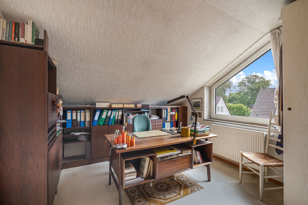 Arbeitszimmer mit Schreibtisch, Regalen, Aktenordnern und großem Fenster unter einer Dachschräge.