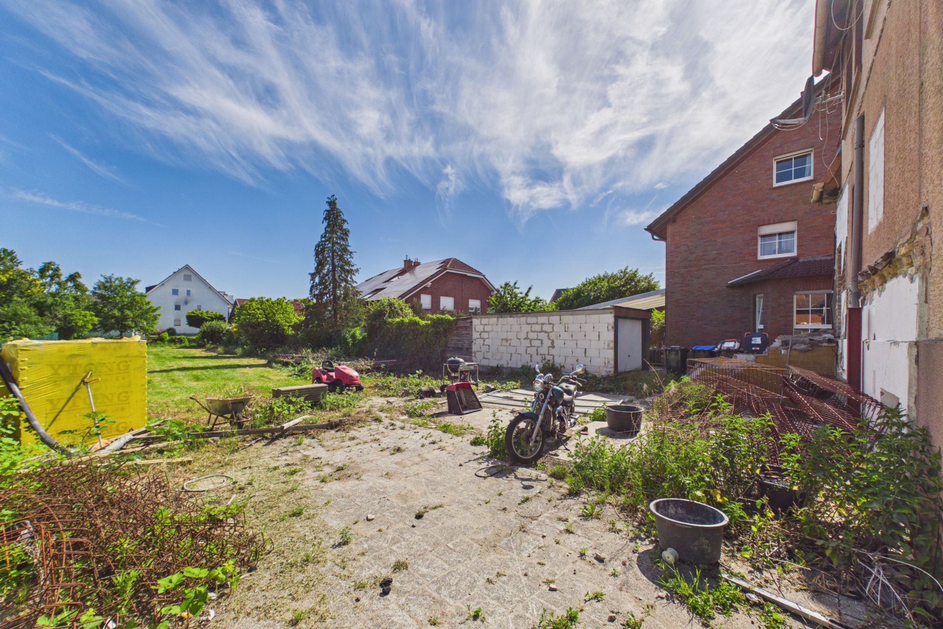 Gepflasterter Hinterhof mit Motorrad, Pflanzen und Geräten neben einem Wohnhaus.