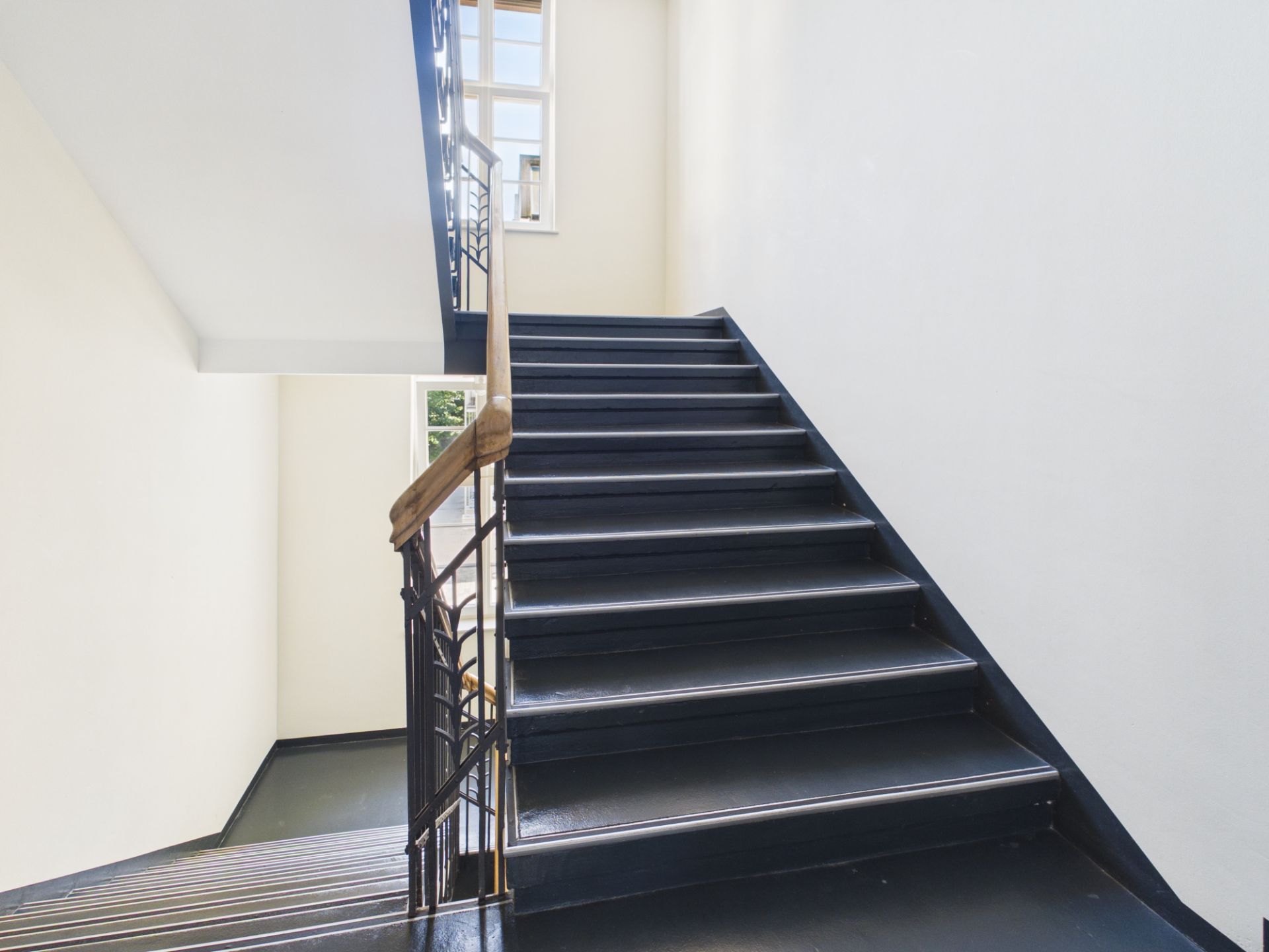 Treppenhaus mit schwarzer Treppe, Geländer aus Metall und Holz sowie großen Fenstern.