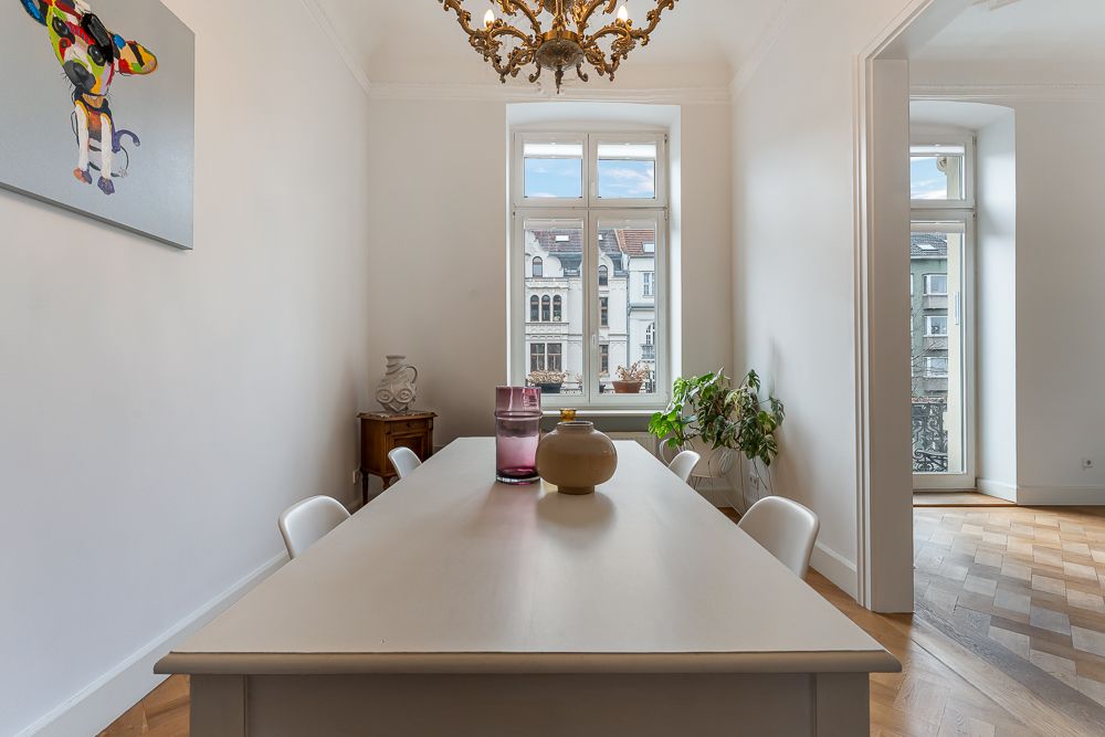 Helles Esszimmer mit weißem Tisch, Stühlen, Kronleuchter und Fenster mit Blick nach draußen.