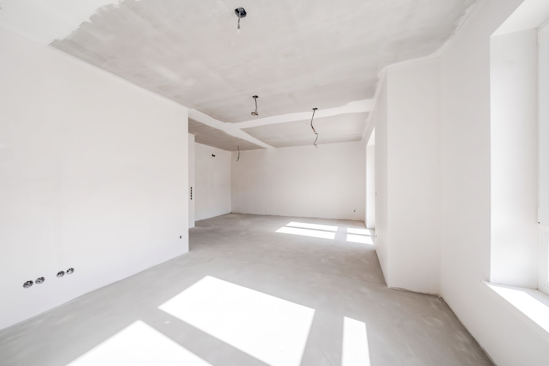 Heller, leerer Raum mit weißen Wänden, großen Fenstern und Kabeln an der Decke.
