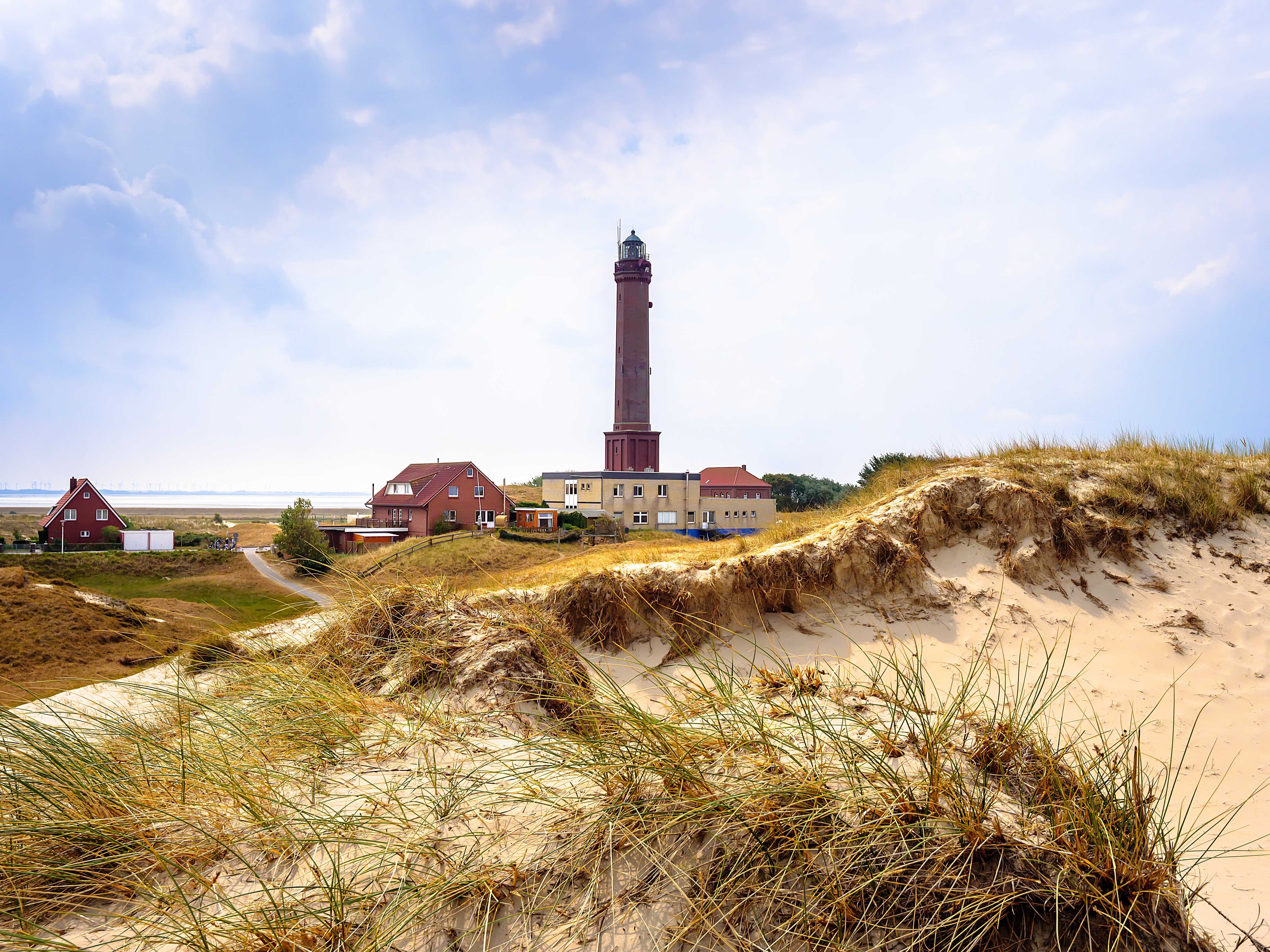 Leuchtturm und Wohnhäuser hinter Sanddünen mit Gras unter bewölktem Himmel.