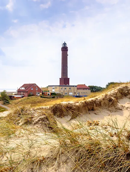 Leuchtturm und Wohnhäuser hinter Sanddünen mit Gras unter bewölktem Himmel.