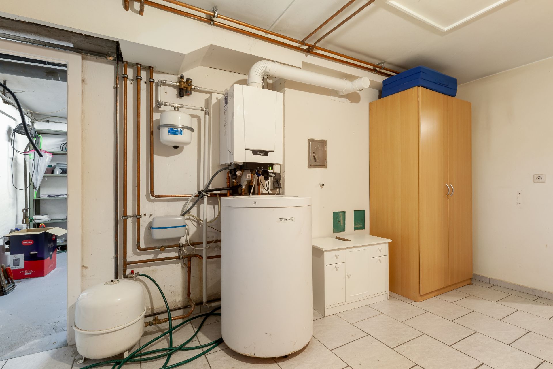 Heizungsraum mit Kessel, Rohren, kleinem Schrank und Holzschrank, gefliestem Boden.
