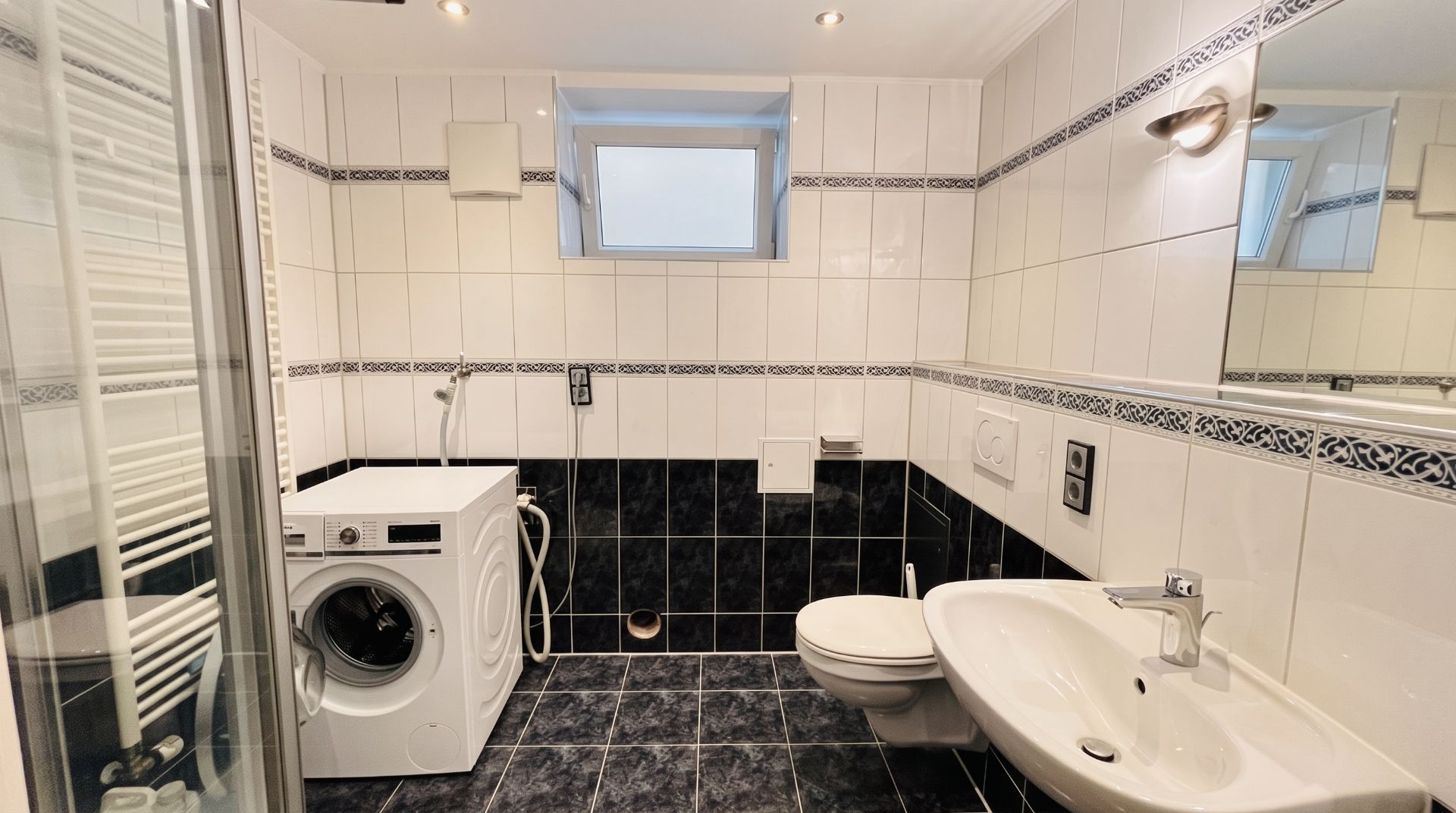 Modernes Bad mit Waschmaschine, Wandtoilette, Waschbecken und Fenster.