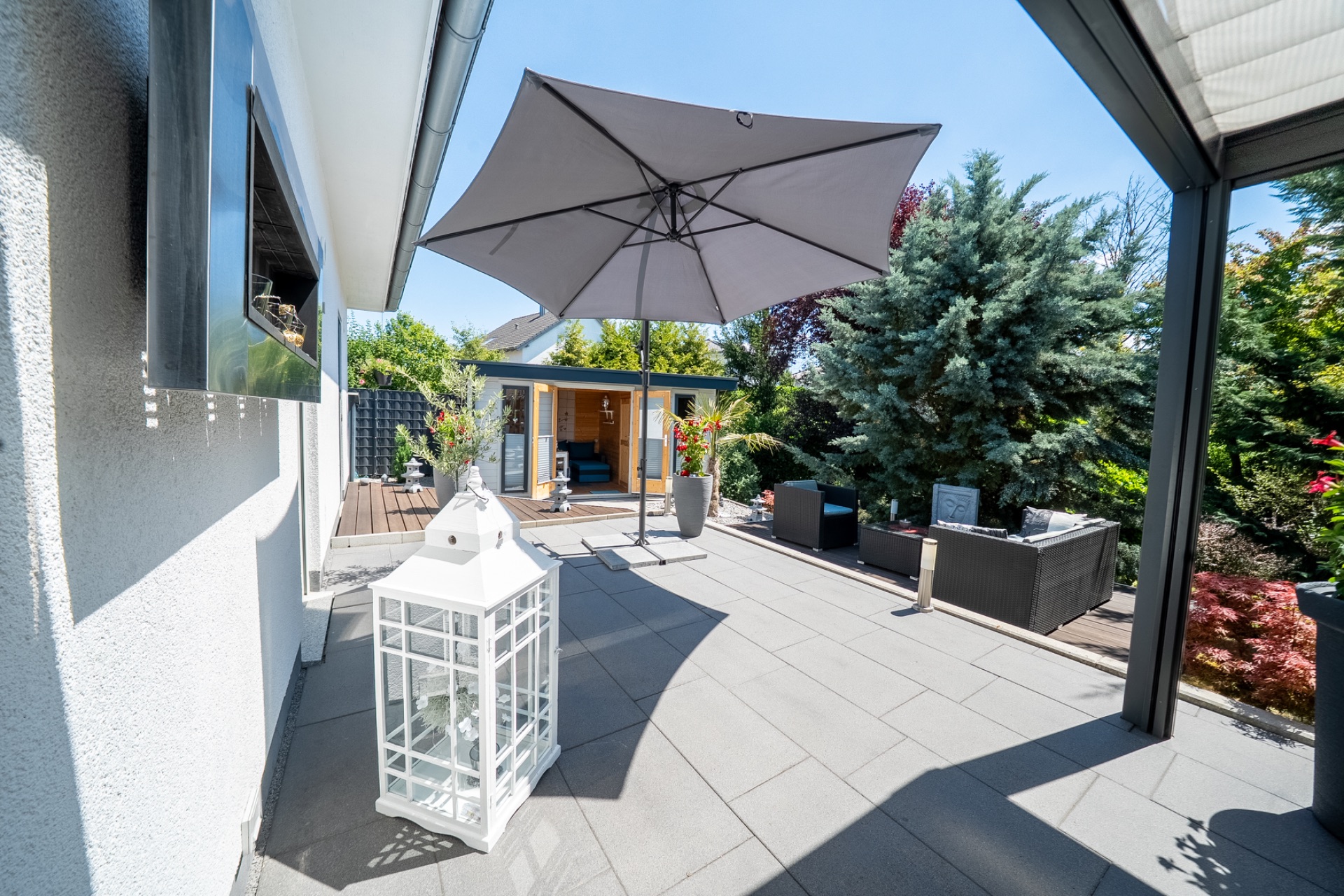 Große Terrasse mit Sonnenschirm, Laterne und Gartenmöbeln, umgeben von Pflanzen.