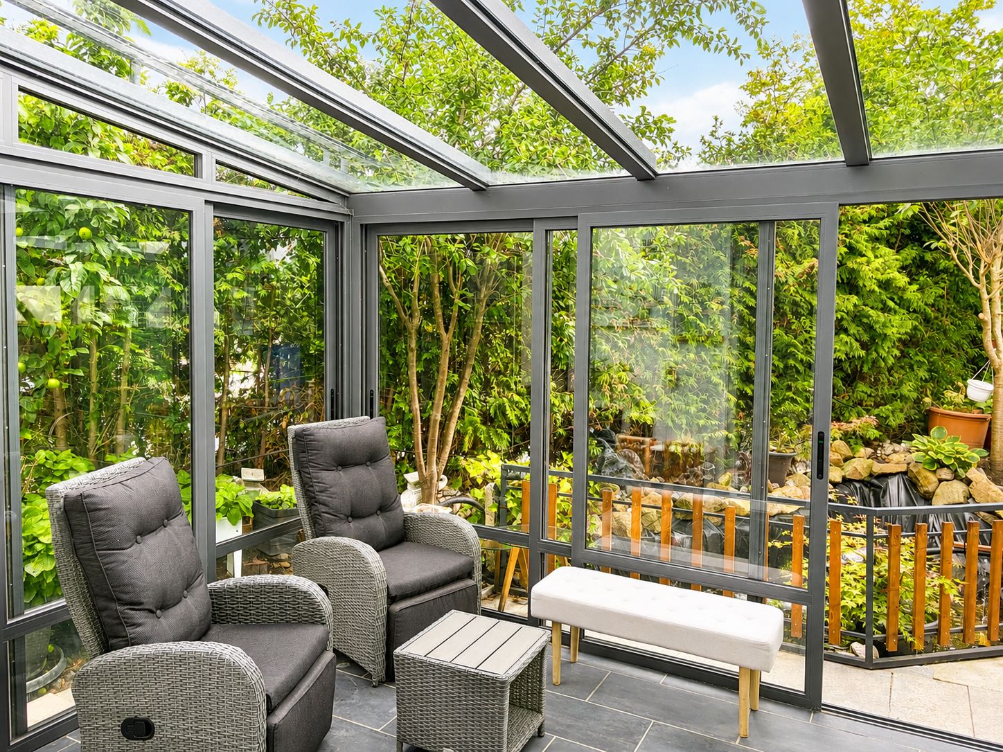 Wintergarten mit zwei grauen Sesseln, kleinem Tisch und Bank, umgeben von viel Grün.