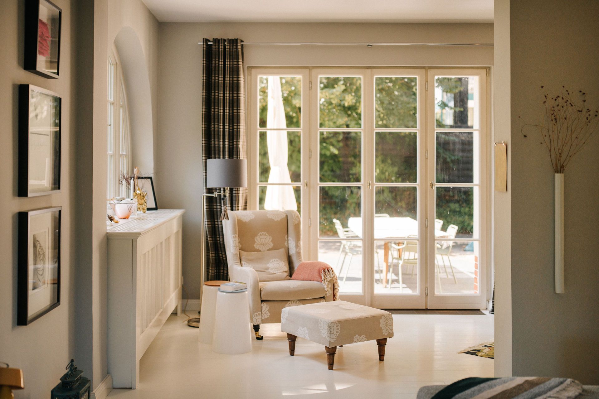 Helles Wohnzimmer mit Sessel, Hocker, Stehlampe und großen Fenstertüren zum Garten.