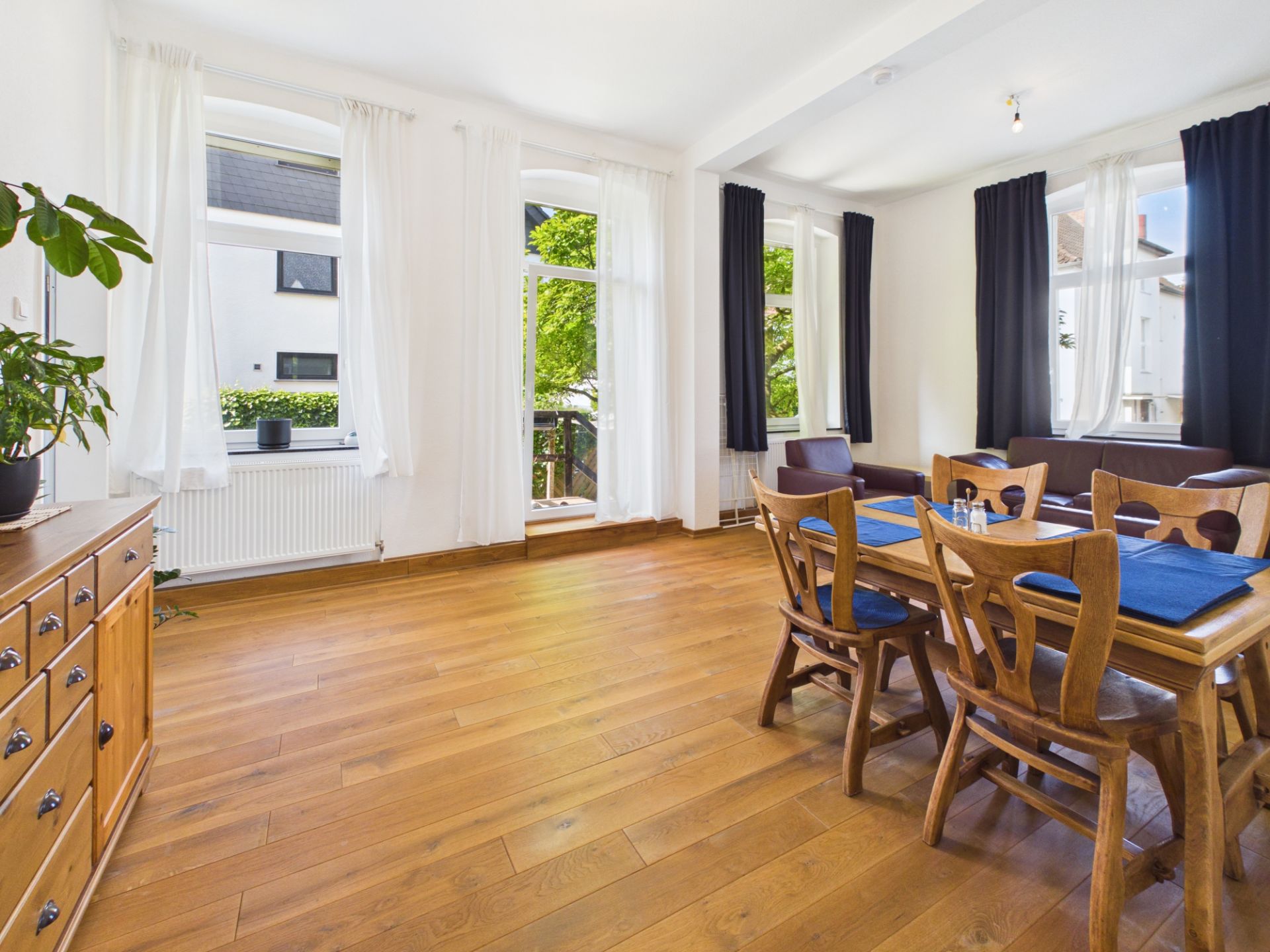 Helles Esszimmer mit Holzboden, großem Tisch, Stühlen und Fenstern mit weißen und dunklen Vorhängen.