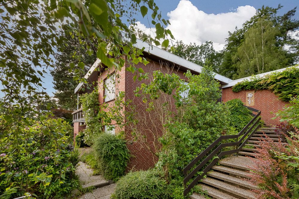 Backsteinhaus mit Flachdach und vielen Pflanzen umgeben, Treppe führt zum Eingang.