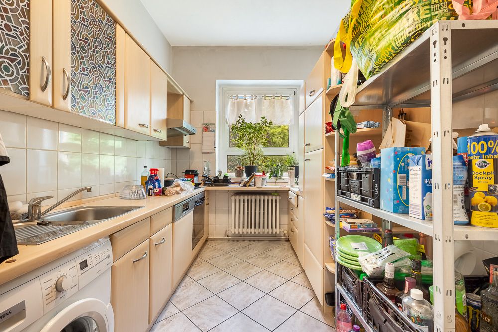 Kleine Küche mit Schränken, Regalen, Spüle, Waschmaschine und Fenster mit Pflanzen.