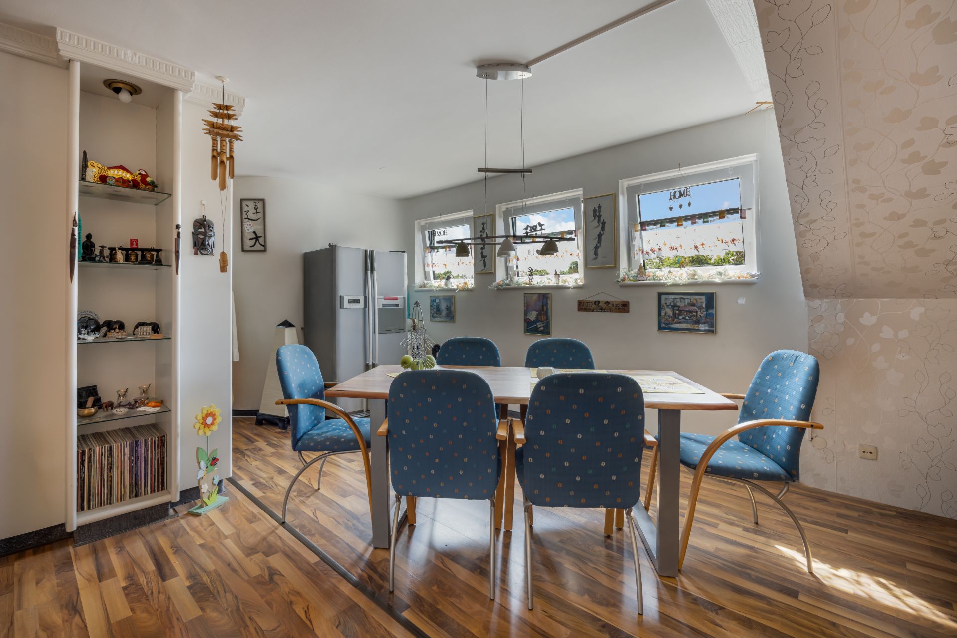 Esszimmer mit Holzboden, rechteckigem Tisch und sechs blauen Stühlen vor drei Fenstern.