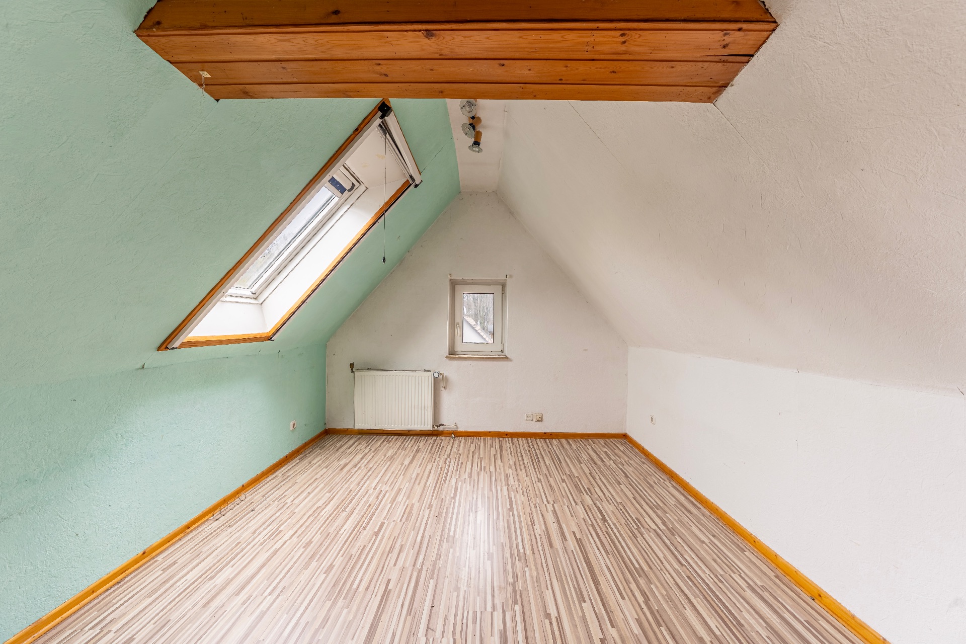 Heller Dachraum mit schrägem Fenster, Laminatboden und kleiner Heizung unter einem Fenster.