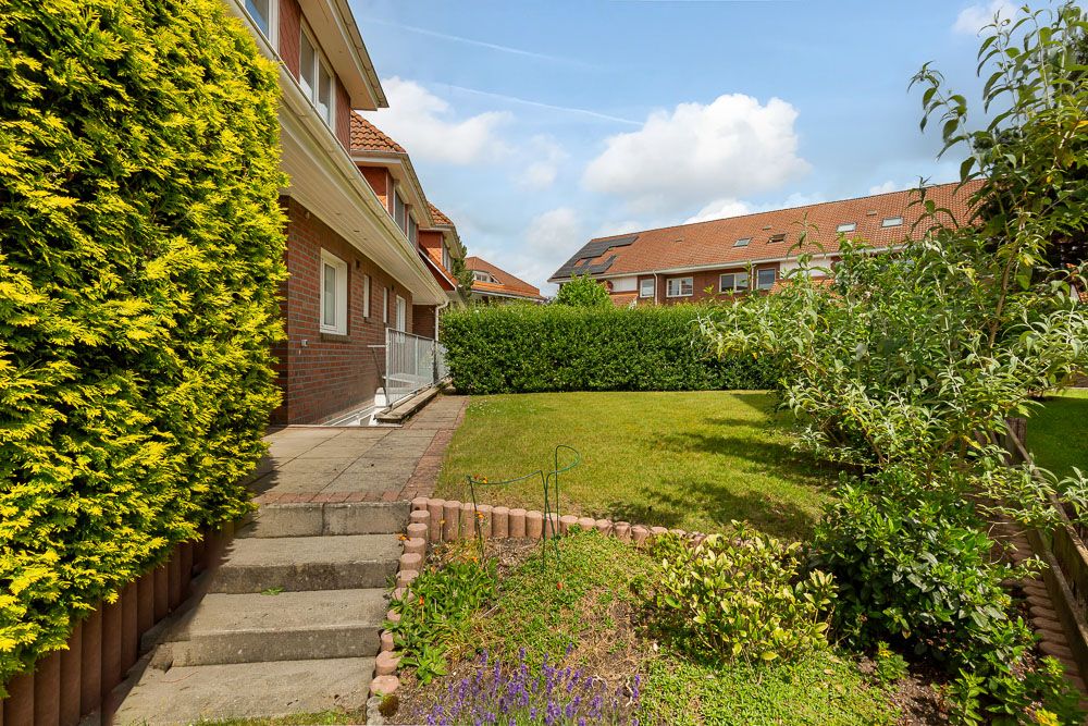 Gepflegter Garten mit Rasen, Terrasse und Büschen vor einem Wohnhaus bei sonnigem Wetter.