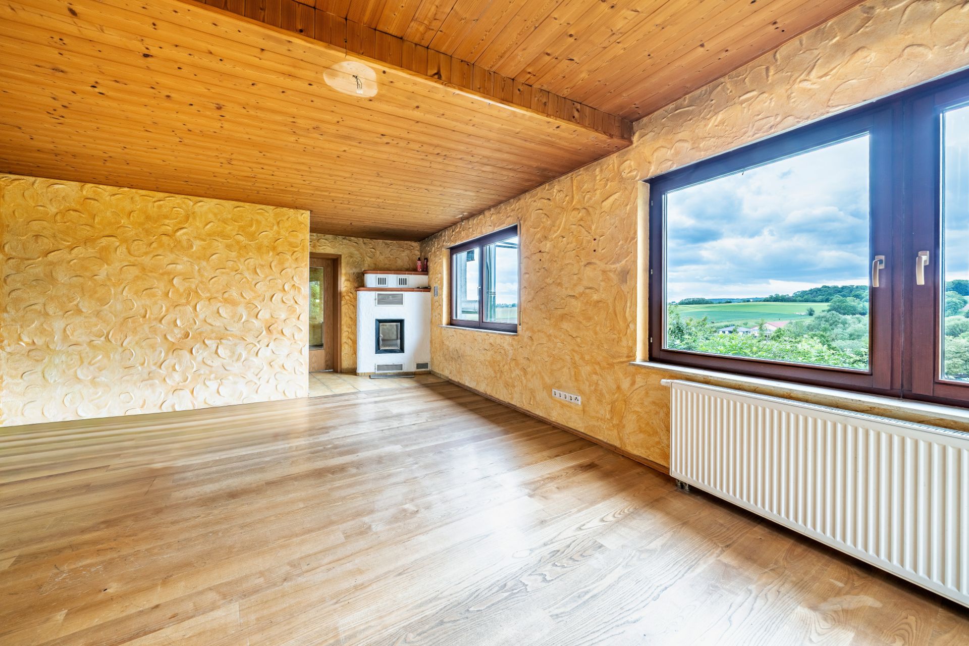 Helles Wohnzimmer mit Holzdecke, großen Fenstern, Parkettboden und Blick ins Grüne.