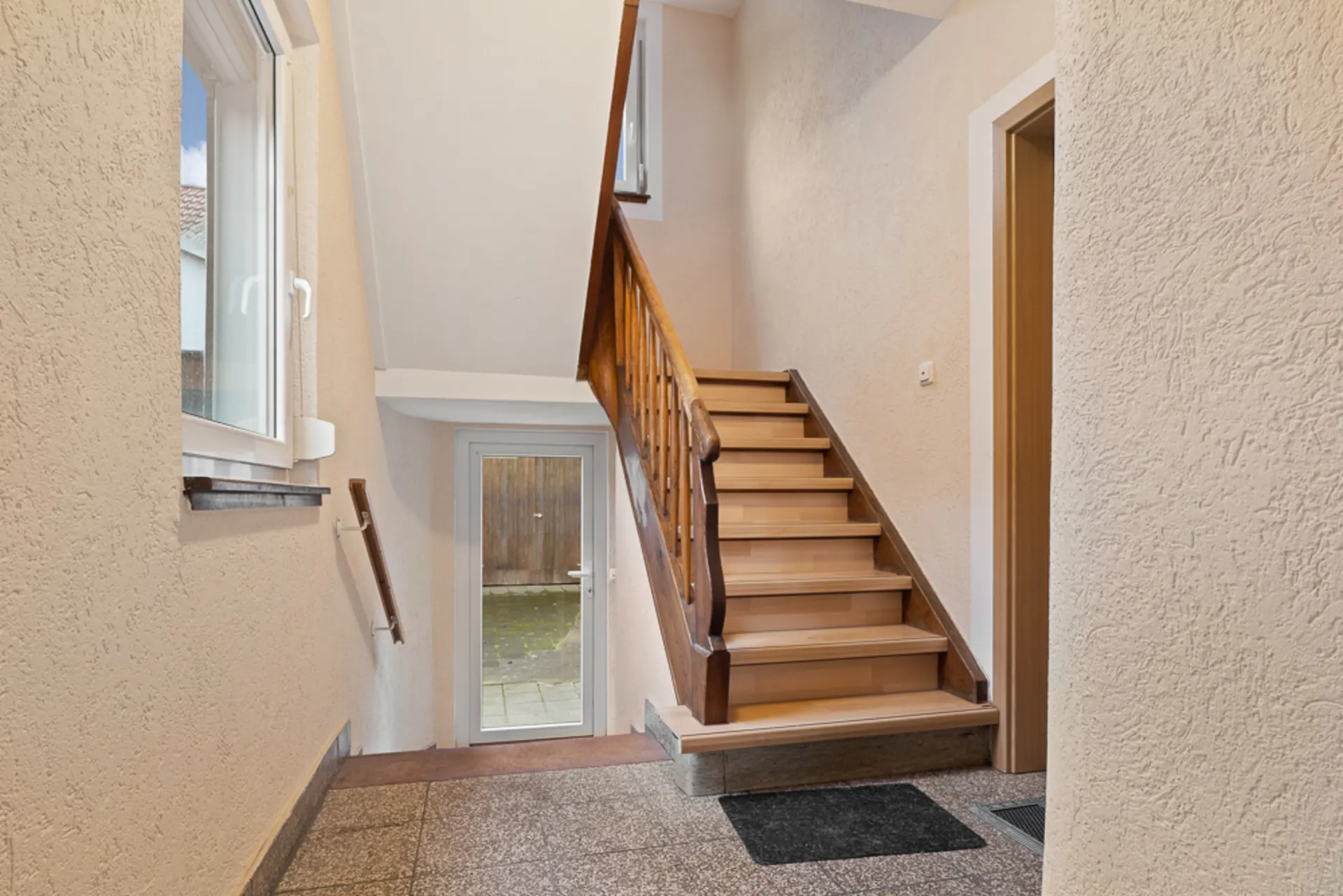 Heller Treppenhausbereich mit Holztreppe, Fenster und Glastür nach draußen.