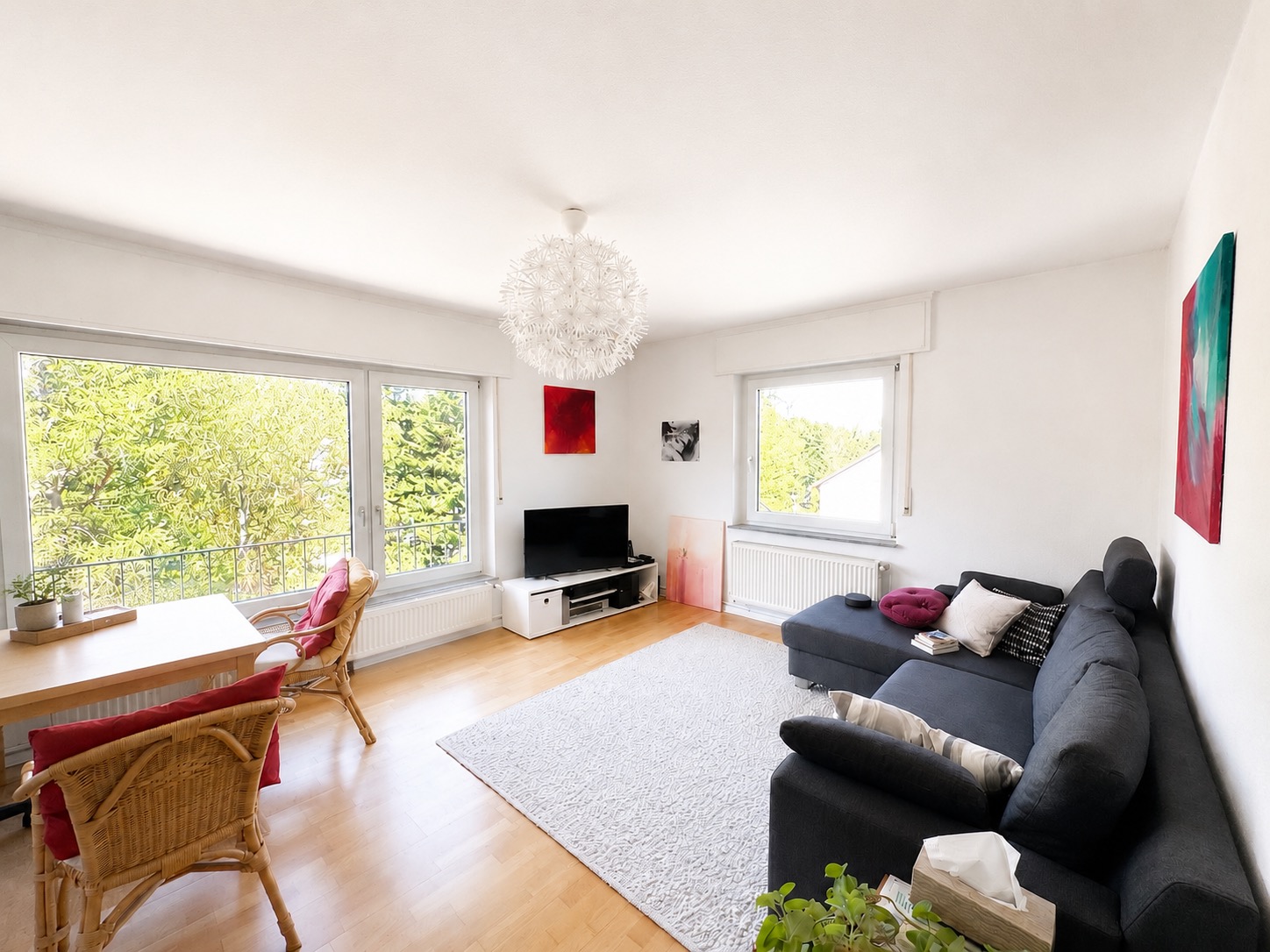 Helles Wohnzimmer mit großem Sofa, Esstisch, Fernseher und großen Fenstern mit Blick ins Grüne.