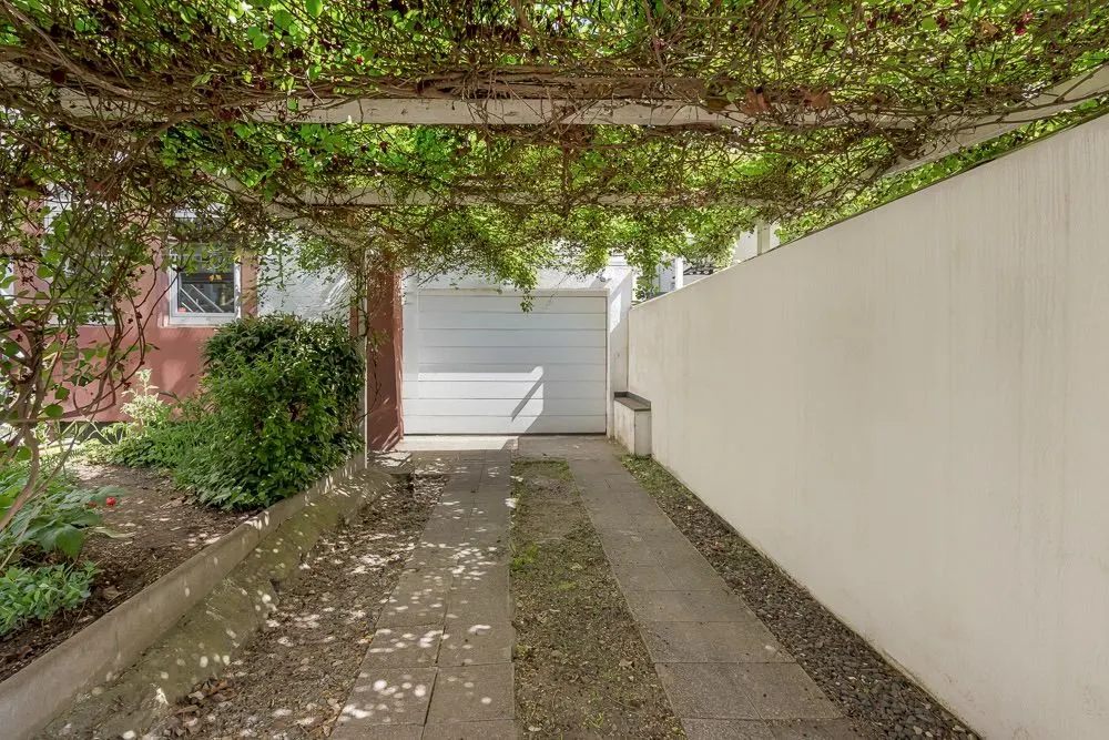 Überdachter Weg mit Pflanzen, führt zu einer weißen Garagentür, daneben Garten und weiße Mauer.
