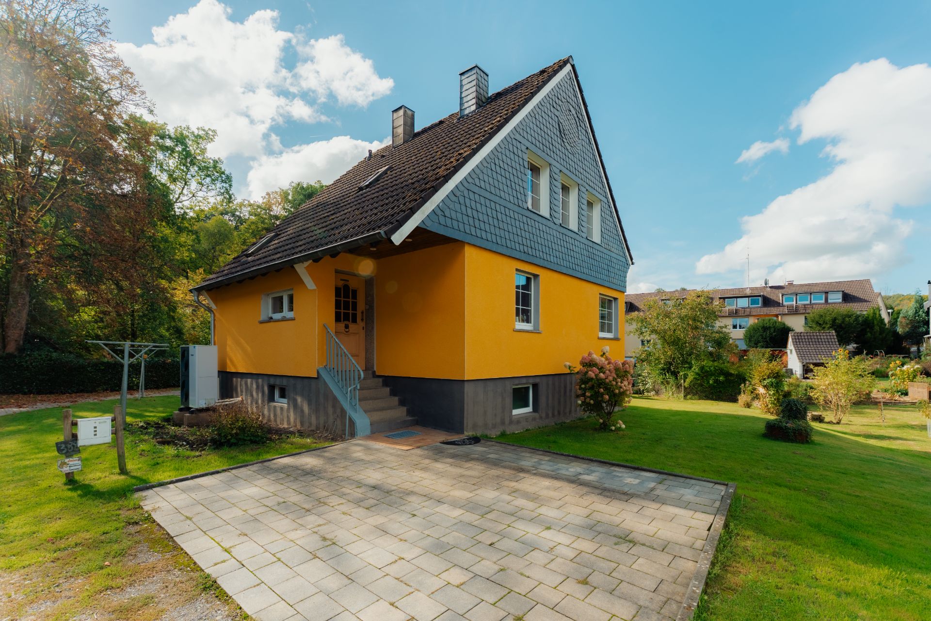 Gelbes Einfamilienhaus mit dunklem Dach, kleiner Treppe und gepflasterter Einfahrt im Garten.