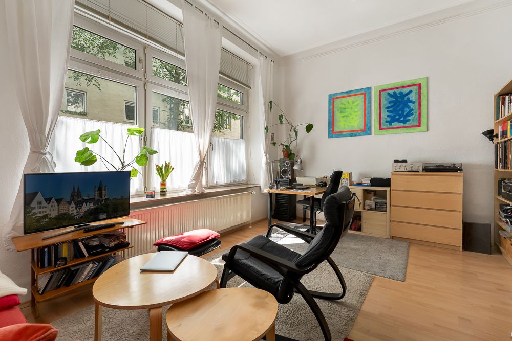 Helles Wohnzimmer mit großen Fenstern, Sessel, Fernseher, Pflanzen und Regalen.