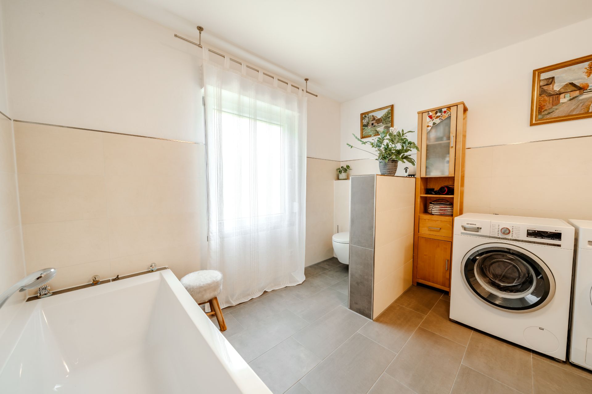 Helles Badezimmer mit Badewanne, Waschmaschine, Holzschrank und WC, Tageslicht durch Fenster mit Vorhang.