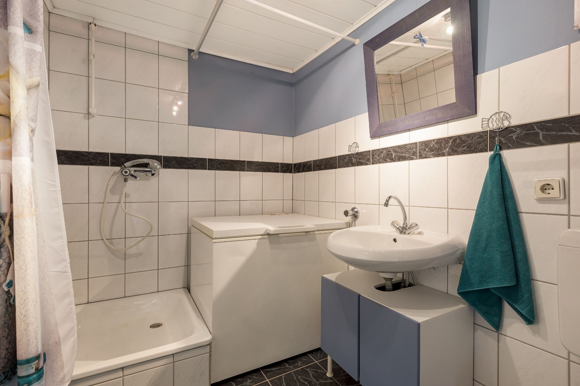 Kleines Bad mit Dusche, Waschbecken, Spiegel, Gefriertruhe und türkisfarbenem Handtuch.