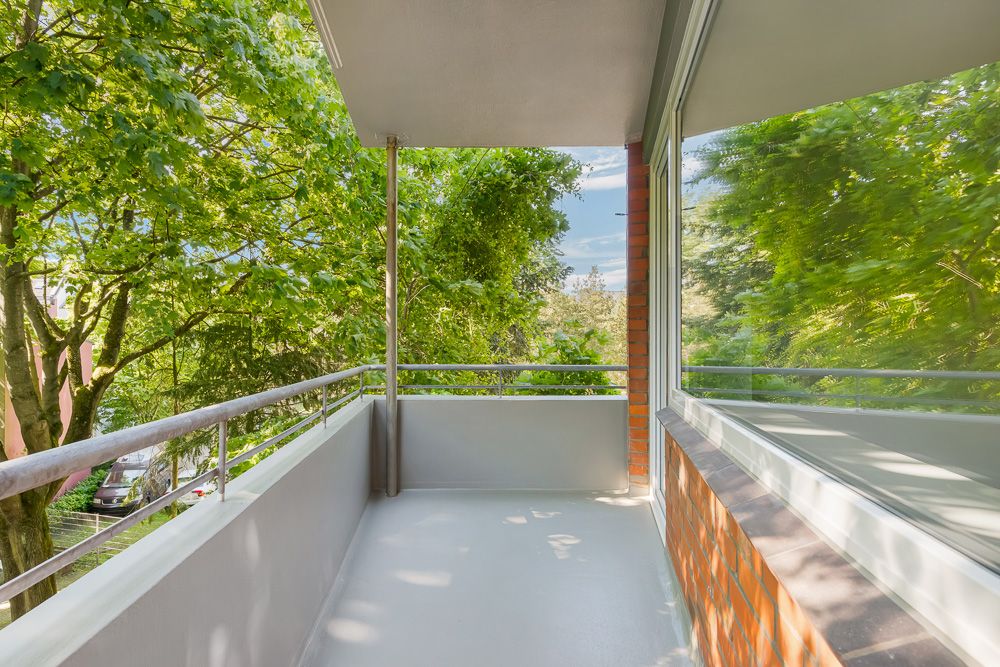 Ein leerer Balkon mit Geländer und Blick auf grüne Bäume, rechts eine große Fensterfront.