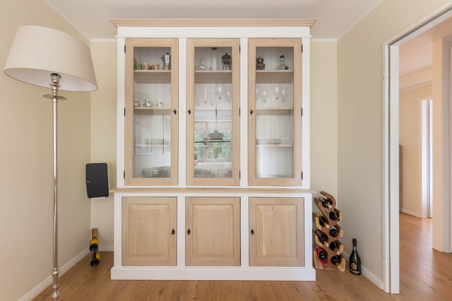 Holzschrank mit Glastüren, daneben ein Weinständer und eine Stehlampe in einem hellen Raum.