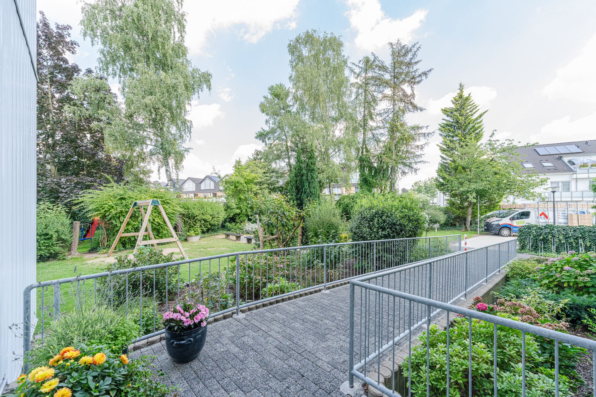 Garten mit Spielplatz, Blumen, Bäumen und Rampe neben Wohnhäusern und geparktem Auto.