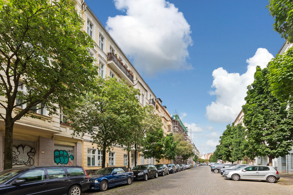 Baumgesäumte Wohnstraße mit parkenden Autos und mehrstöckigen Häusern an einem sonnigen Tag.