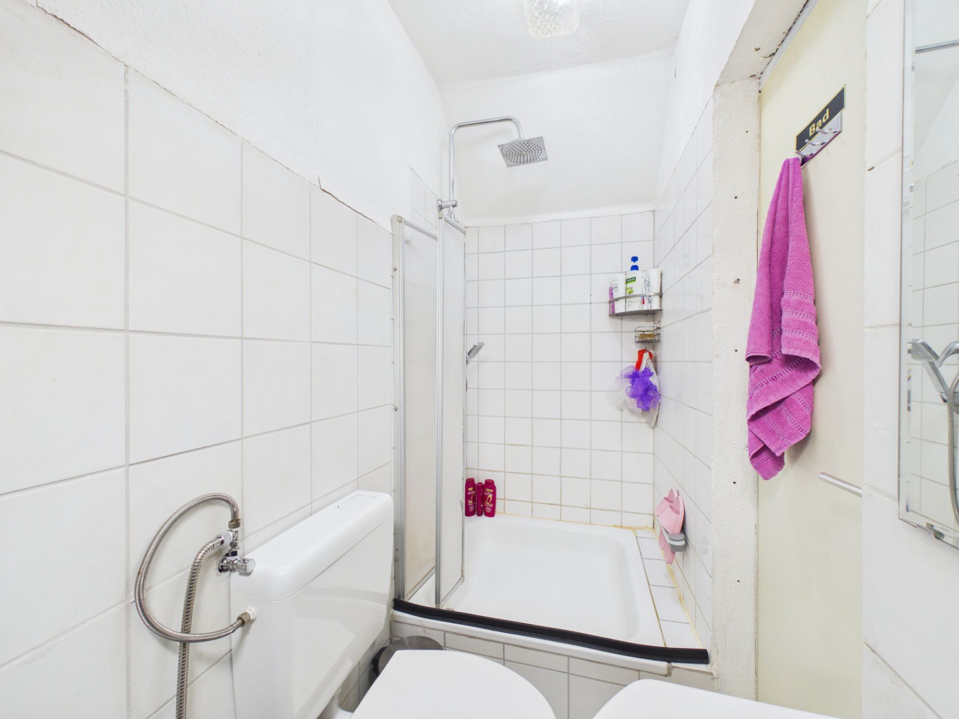 Kleines Badezimmer mit Dusche, Toilette, weißen Fliesen und einem lila Handtuch an der Tür.