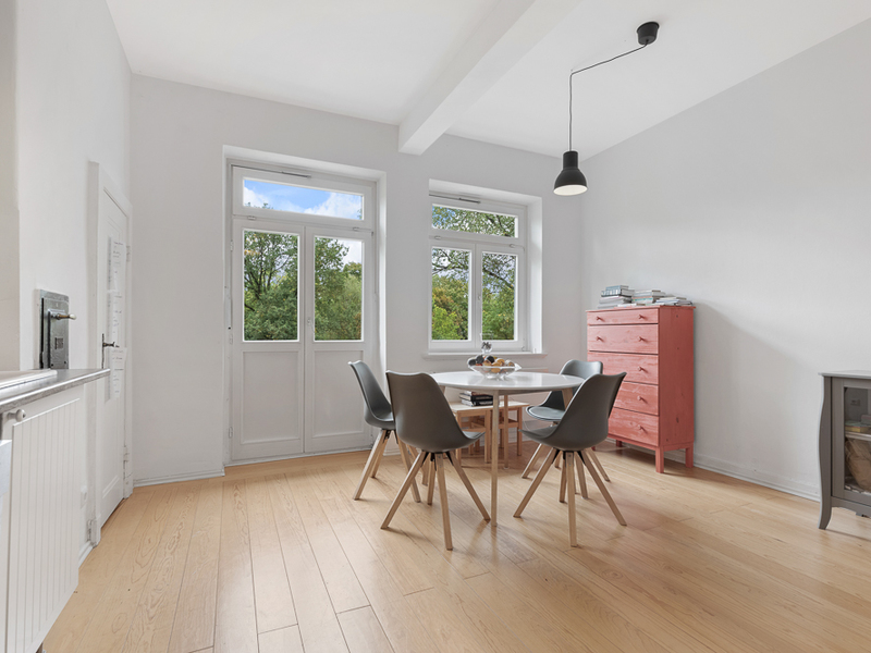 Helles Esszimmer mit rundem Tisch, vier Stühlen und roter Kommode vor großen Fenstern.