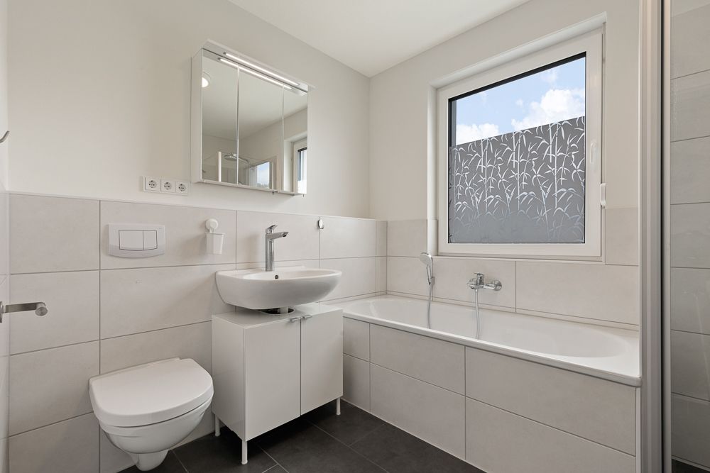 Helles Badezimmer mit Badewanne, Waschbecken, Toilette und Fenster mit Sichtschutzfolie.