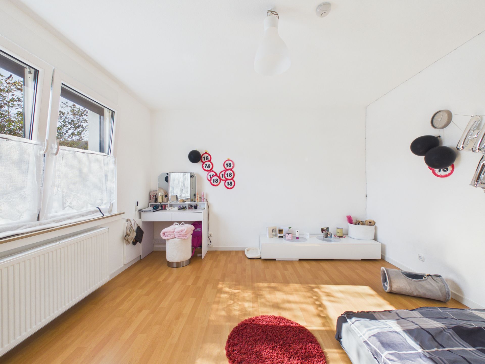 Helles Zimmer mit Holzboden, großem Fenster, Schminktisch und wenigen Möbeln.