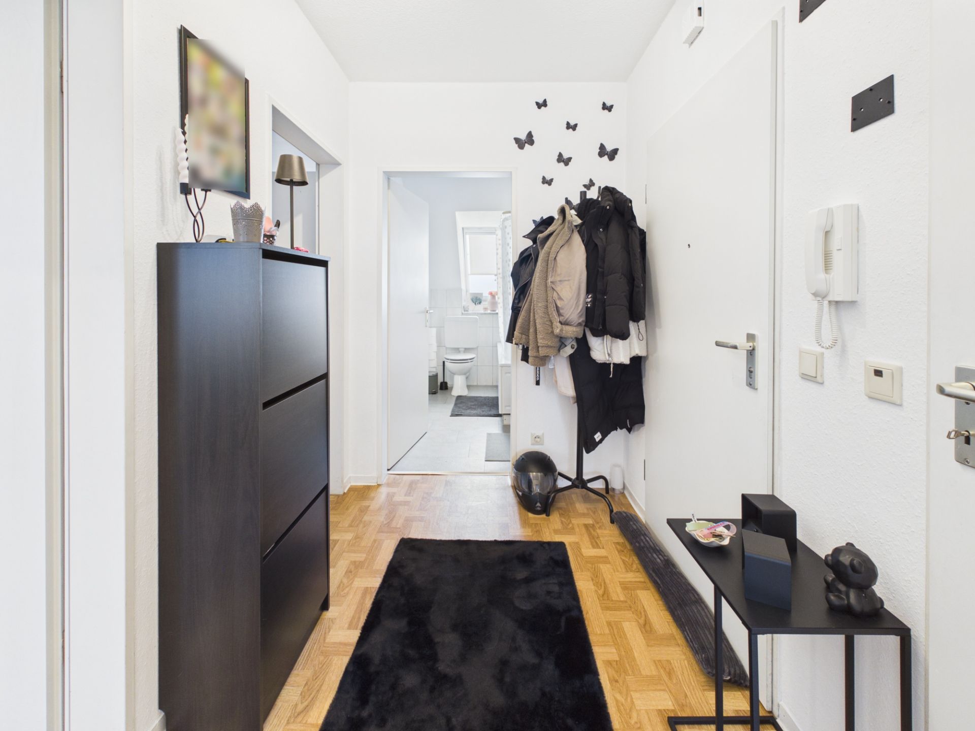 Heller Flur mit Garderobe, schwarzem Teppich, Kommode und Blick ins Badezimmer.